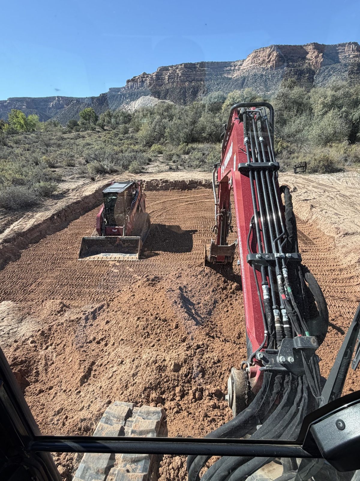 View from the CJC mini excavator cab over a graded site, with a skid steer working below red rock cliffs